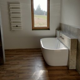 Przemysław Rolnik - Nowoczesna łazienka z wolnostojącą białą wanną, drewnopodobną podłogą, grzejnikiem drabinkowym na ścianie i oknem z widokiem na zielone pole. Widoczne niedokończone prace elektryczne.