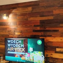 Przemysław Rolnik - Ściana w salonie wykończona panelami z naturalnego drewna o zróżnicowanej kolorystyce, oświetlona lampą ścienną, z widocznym telewizorem i fragmentem otwartego przejścia do innego pomieszczenia.