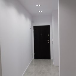 Przemysław Rolnik - Wnętrze korytarza po remoncie z czarnymi drzwiami wejściowymi, białymi ścianami i szarą podłogą, oświetlone punktowymi lampami sufitowymi.