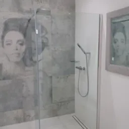 Nowoczesna kabina prysznicowa z szarymi płytkami imitującymi beton, zdobiona portretem kobiety, z odpływem liniowym i lustrem z odbiciem tej samej twarzy.