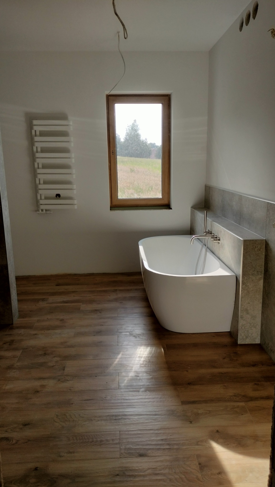Nowoczesna łazienka z wolnostojącą białą wanną, drewnopodobną podłogą, grzejnikiem drabinkowym na ścianie i oknem z widokiem na zielone pole. Widoczne niedokończone prace elektryczne.