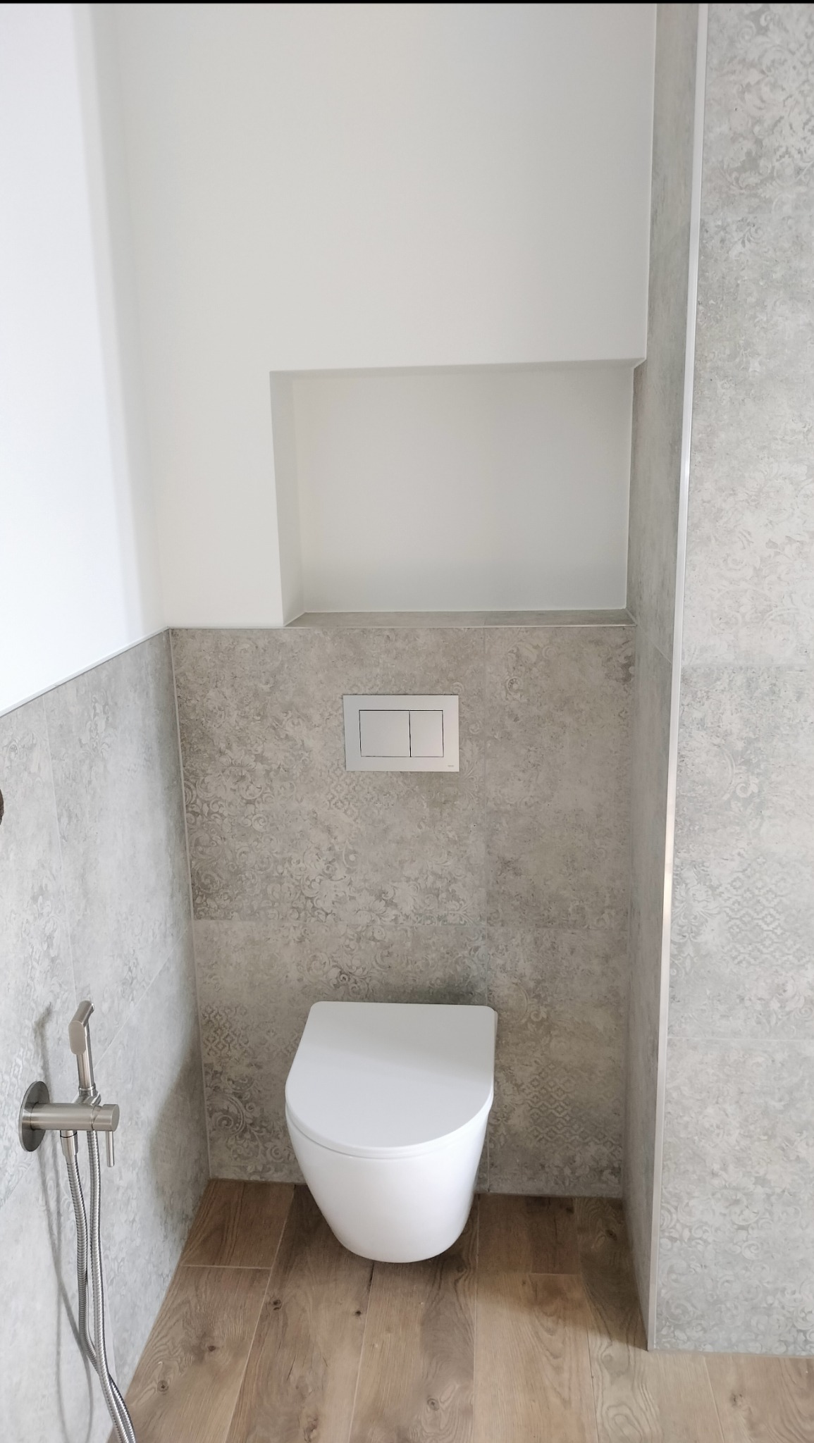 Nowoczesna toaleta podwieszana z szarymi płytkami w marokański wzór, drewnopodobna podłoga i bateria bidetowa ze stali nierdzewnej.