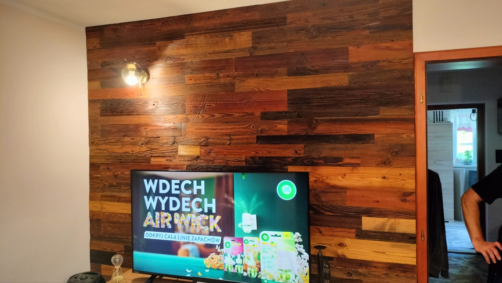 Ściana w salonie wykończona panelami z naturalnego drewna o zróżnicowanej kolorystyce, oświetlona lampą ścienną, z widocznym telewizorem i fragmentem otwartego przejścia do innego pomieszczenia.