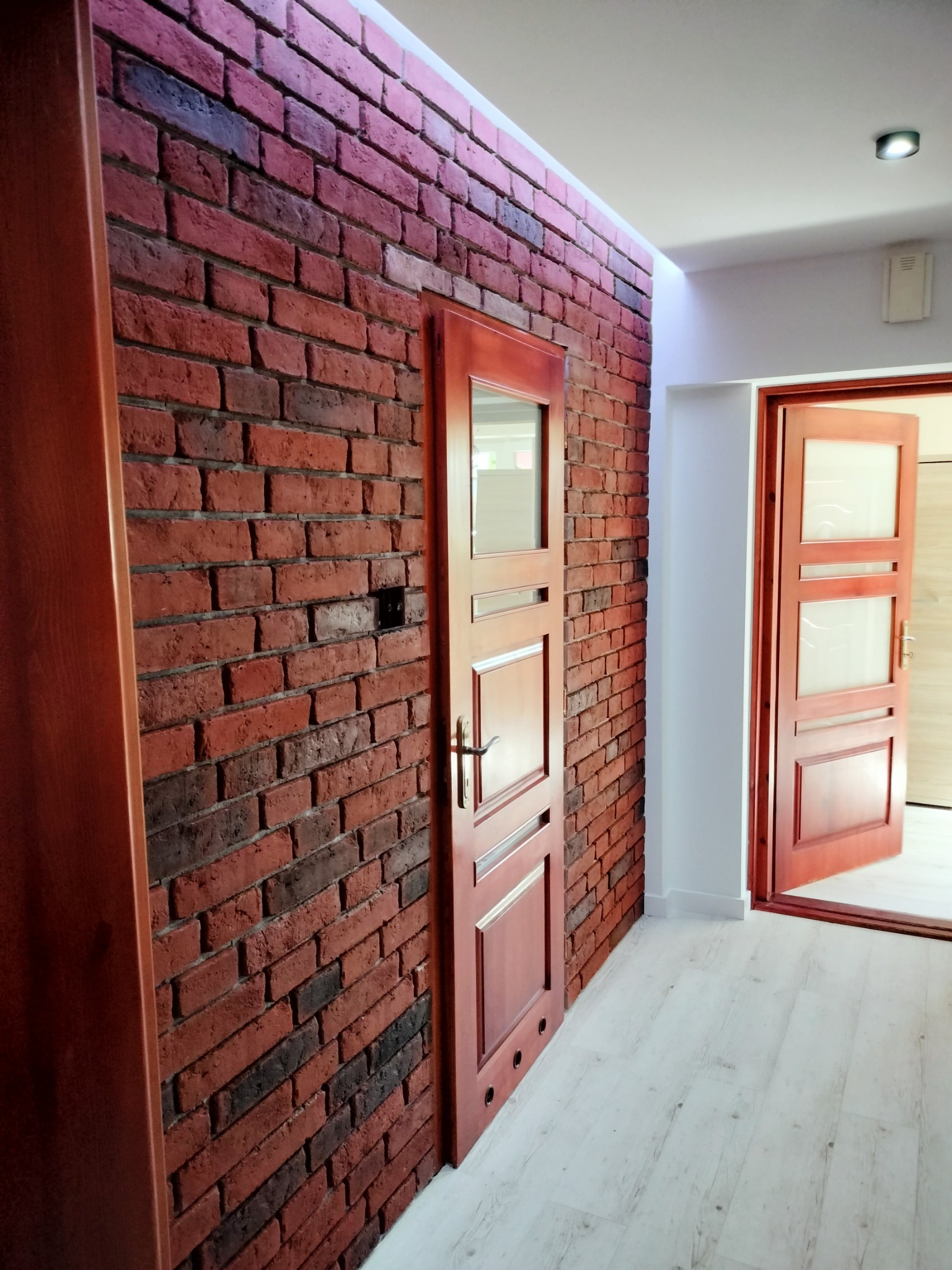 Ceglana ściana z czerwonobrązowych cegieł z widoczną instalacją elektryczną, drewniane drzwi wewnętrzne z przeszkleniami, biała podłoga i oświetlenie sufitowe.