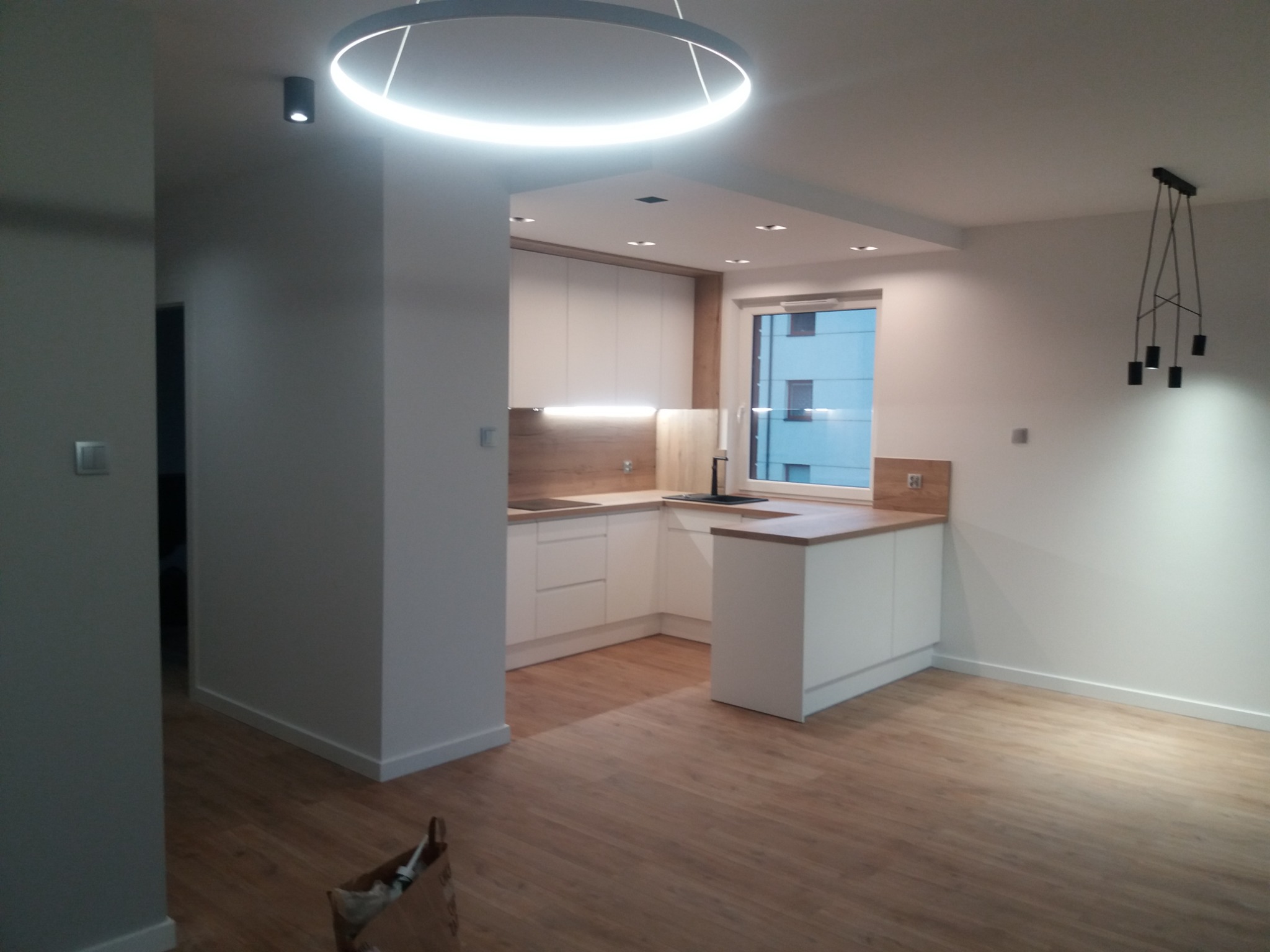 Nowoczesne wnętrze mieszkania po remoncie: jasna kuchnia z drewnianym blatem, minimalistyczne oświetlenie sufitowe i parkiet w odcieniach brązu, widoczny fragment salonu z designerską lampą wiszącą.