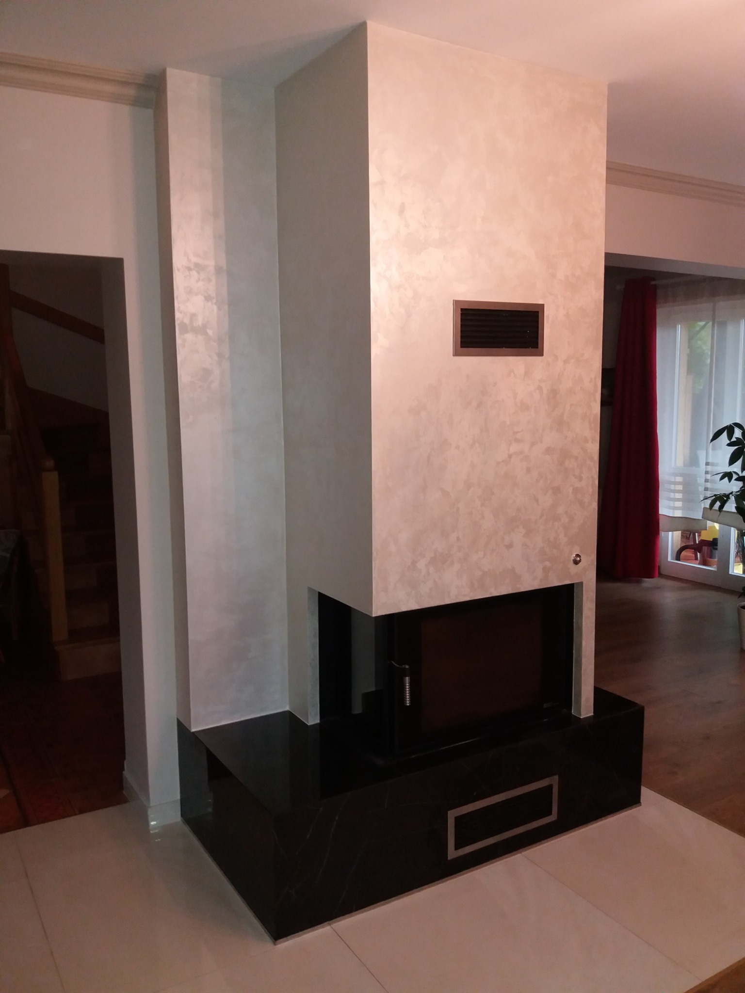 Elegancki kominek z czarną podstawą i wykończeniem w kolorze kremowym z efektem dekoracyjnym na ścianie, widoczny fragment podłogi z jasnych płytek oraz drewniana podłoga w tle, czerwone zasłony...