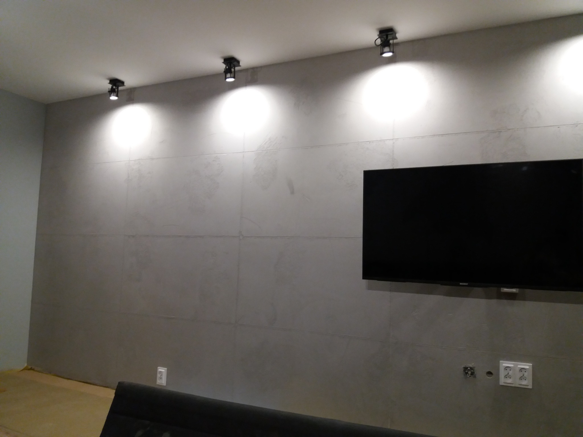 Szara ściana wykończona panelami, z zamontowanym telewizorem i oświetleniem punktowym na suficie; widoczne gniazdka elektryczne.