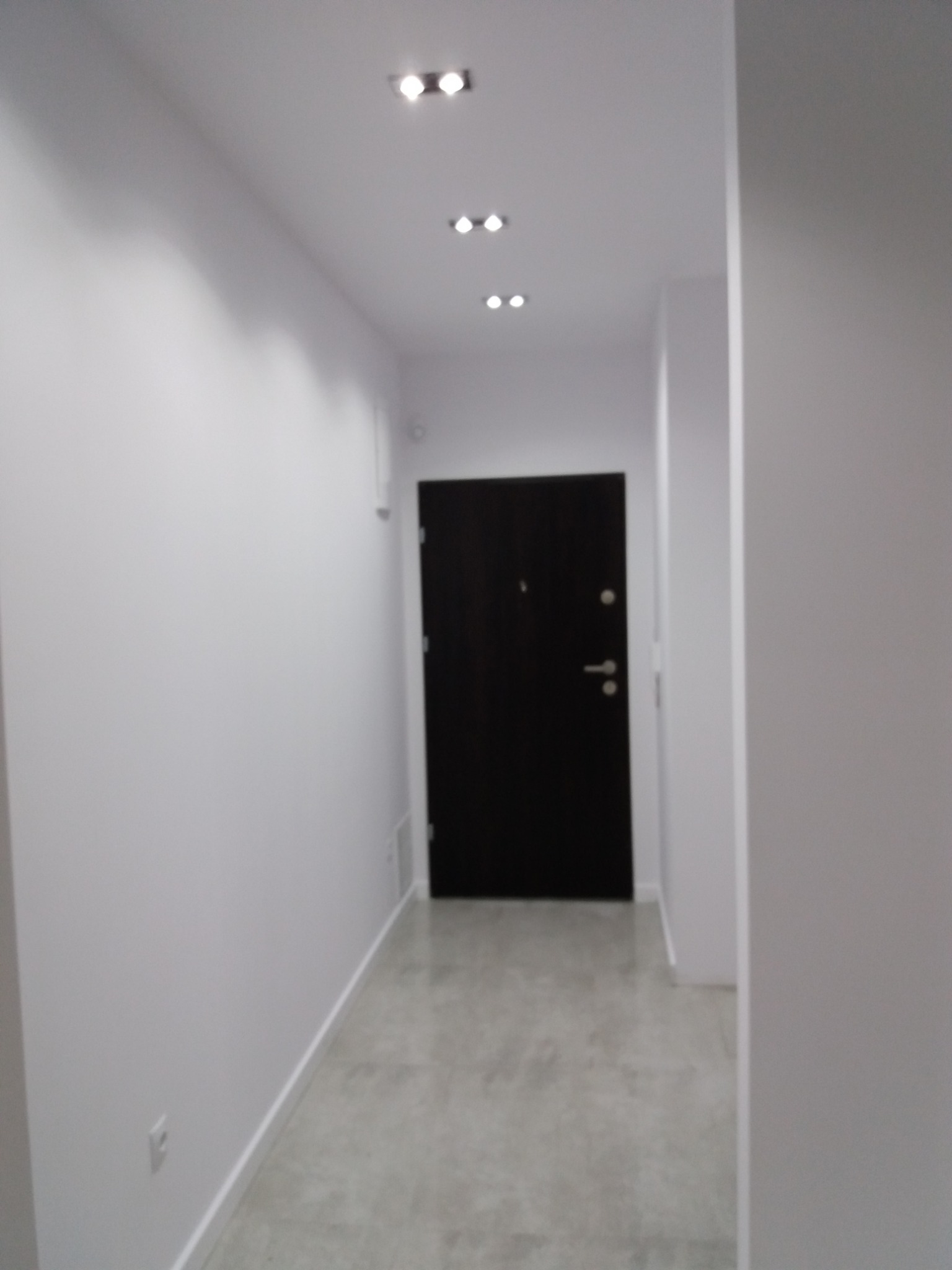 Wnętrze korytarza po remoncie z czarnymi drzwiami wejściowymi, białymi ścianami i szarą podłogą, oświetlone punktowymi lampami sufitowymi.