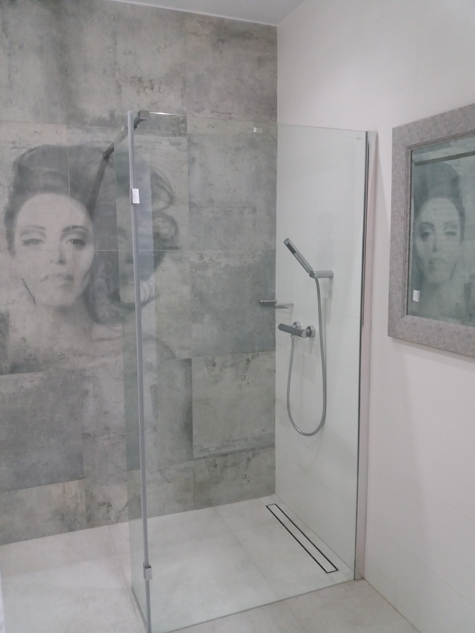 Nowoczesna kabina prysznicowa z szarymi płytkami imitującymi beton, zdobiona portretem kobiety, z odpływem liniowym i lustrem z odbiciem tej samej twarzy.