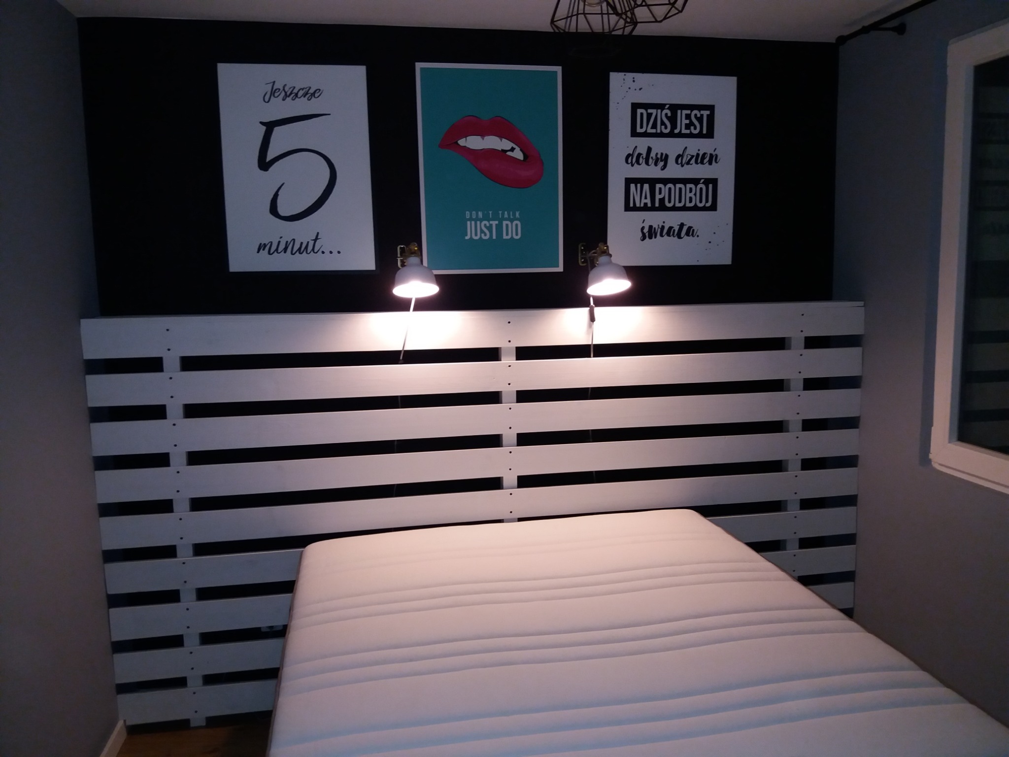 Sypialnia z łóżkiem z palet, czarną ścianą z trzema plakatami i lampkami nocnymi, minimalistyczny design.
