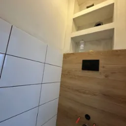 Wnęka z półkami w łazience, częściowo wykończona białymi płytkami i panelami drewnopodobnymi, widoczne otwory instalacyjne i czarny panel spłuczki, butelka wody w jednej z półek.