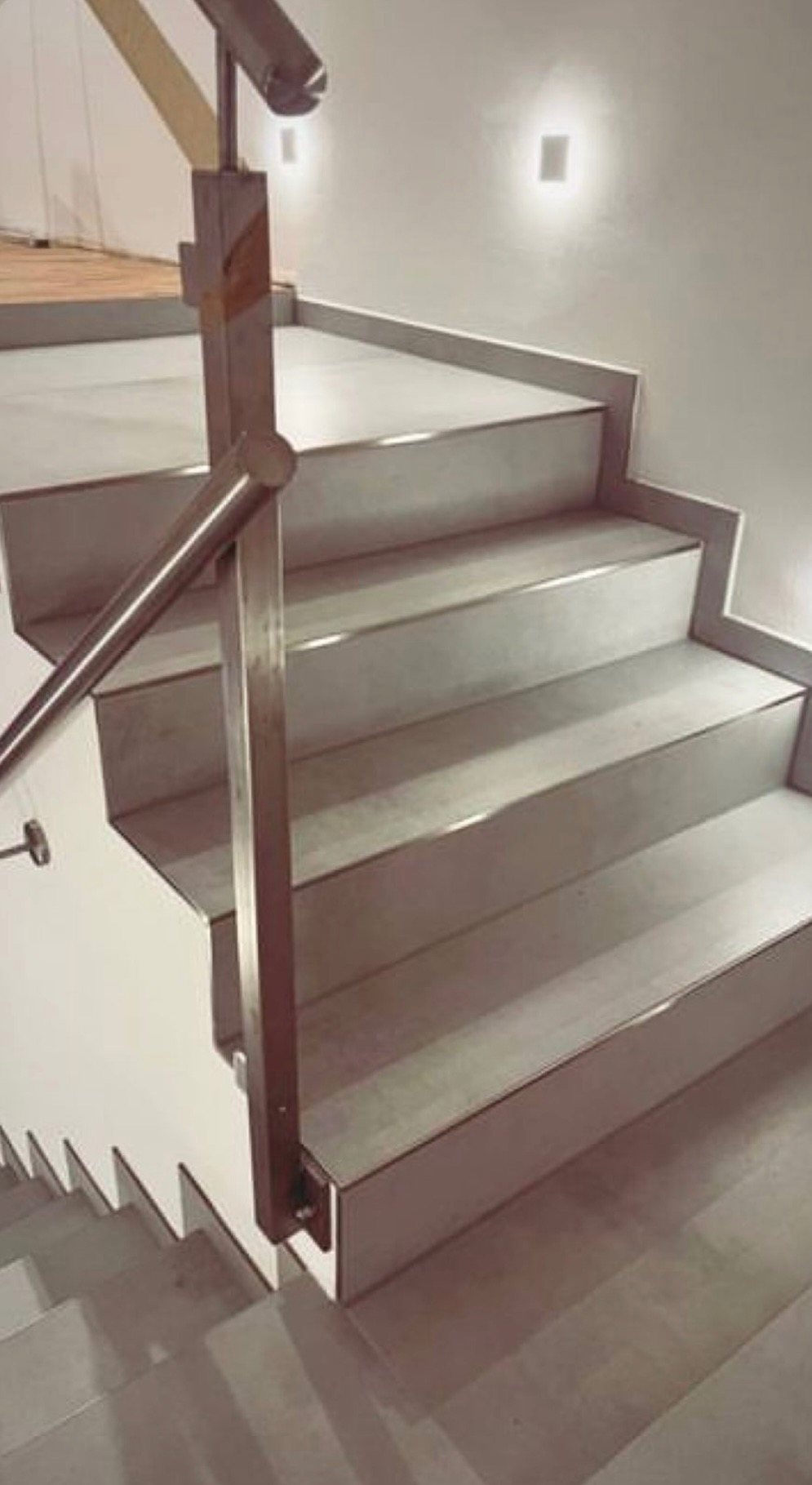 Schody wewnętrzne wykończone płytkami w odcieniach szarości z metalowymi listwami, oświetlenie punktowe na ścianie i chromowana balustrada