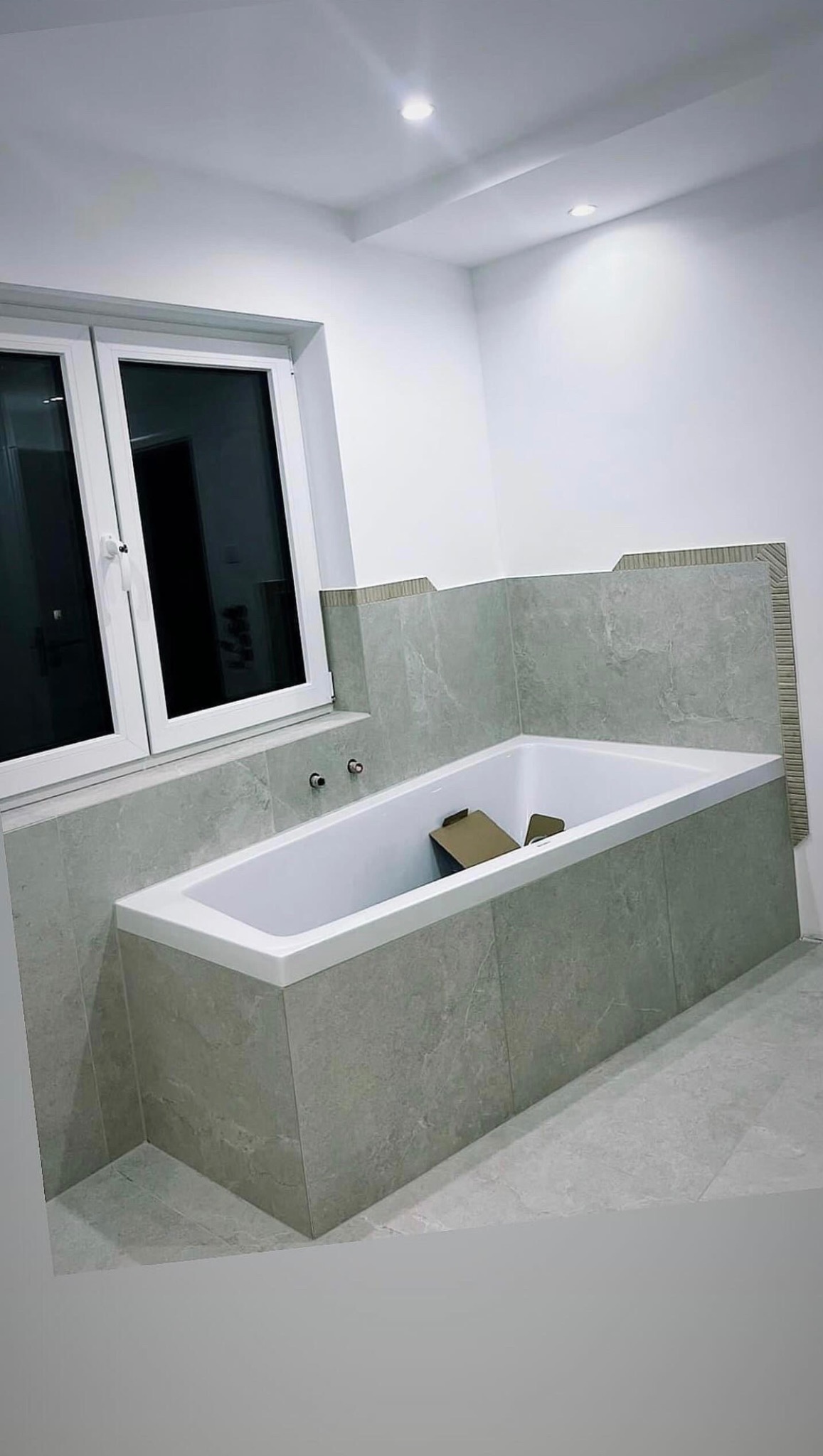 Nowoczesna łazienka z zabudowaną wanną w szarych płytkach imitujących kamień, biała rama okienna i sufit z oświetleniem punktowym, widoczne fragmenty kartonowego opakowania w wannie.