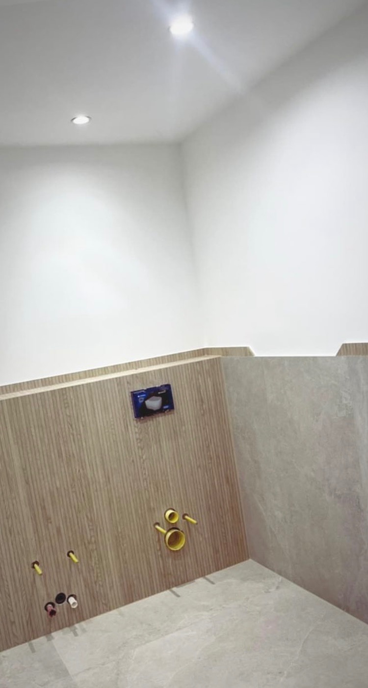 Przygotowana ściana w łazience z wyprowadzonymi instalacjami wodno-kanalizacyjnymi, częściowo wykończona płytkami imitującymi drewno i szarym gresem, widoczna podtynkowa spłuczka WC.