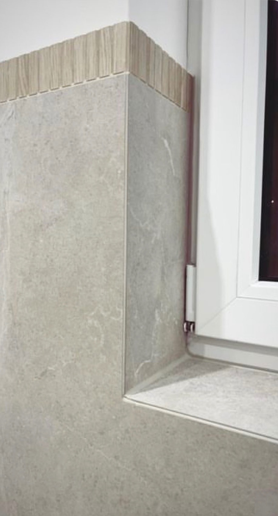 Wykończona wnęka okienna z szarą płytką imitującą kamień i dekoracyjnym pasem mozaiki drewnopodobnej na górze, widoczny fragment białej ramy okiennej.