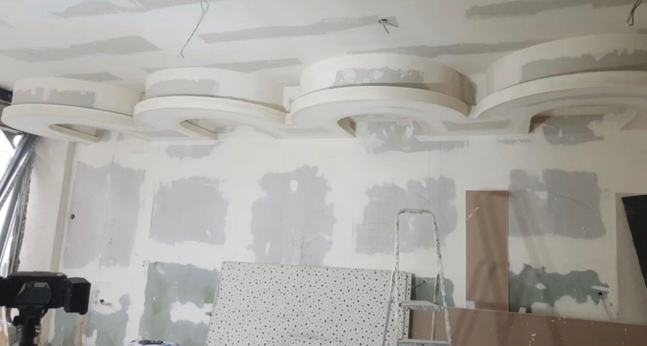 Wnętrze w trakcie prac: dekoracyjny sufit podwieszany z gips-kartonu z półokrągłymi elementami, ściany z widocznymi śladami szpachlowania, drabina i płyta gipsowa w pomieszczeniu.