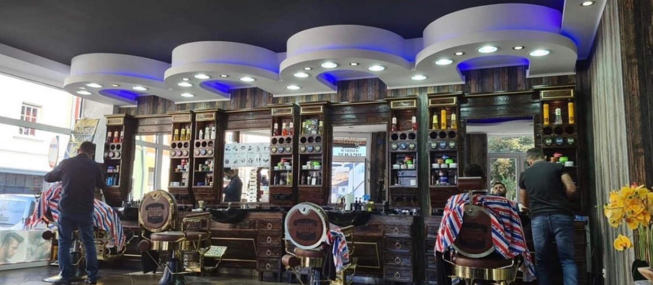 Wnętrze luksusowego salonu fryzjerskiego w stylu vintage z ciemnego drewna, z oświetleniem LED, trzema stanowiskami pracy i dwoma fryzjerami obsługującymi klientów.