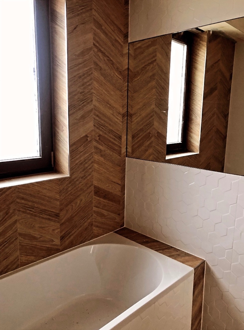 Wykończona łazienka z drewnopodobnymi płytkami ułożonymi w jodełkę na ścianach, białe heksagonalne płytki częściowo odbijające się w lustrze, oraz fragment białej wanny.
