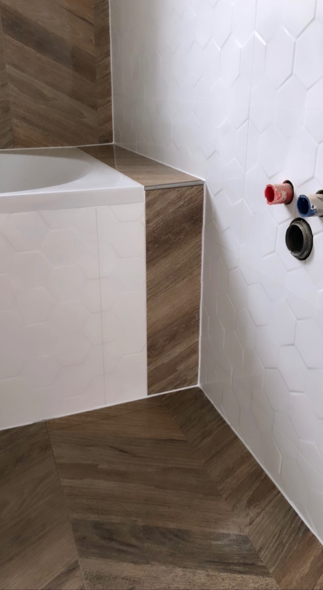 Wykończona łazienka z biało-brązowymi płytkami heksagonalnymi na ścianach, brązowymi płytkami imitującymi drewno na podłodze ułożonymi w jodełkę, obudowa wanny wykończona tymi samymi płytkami...