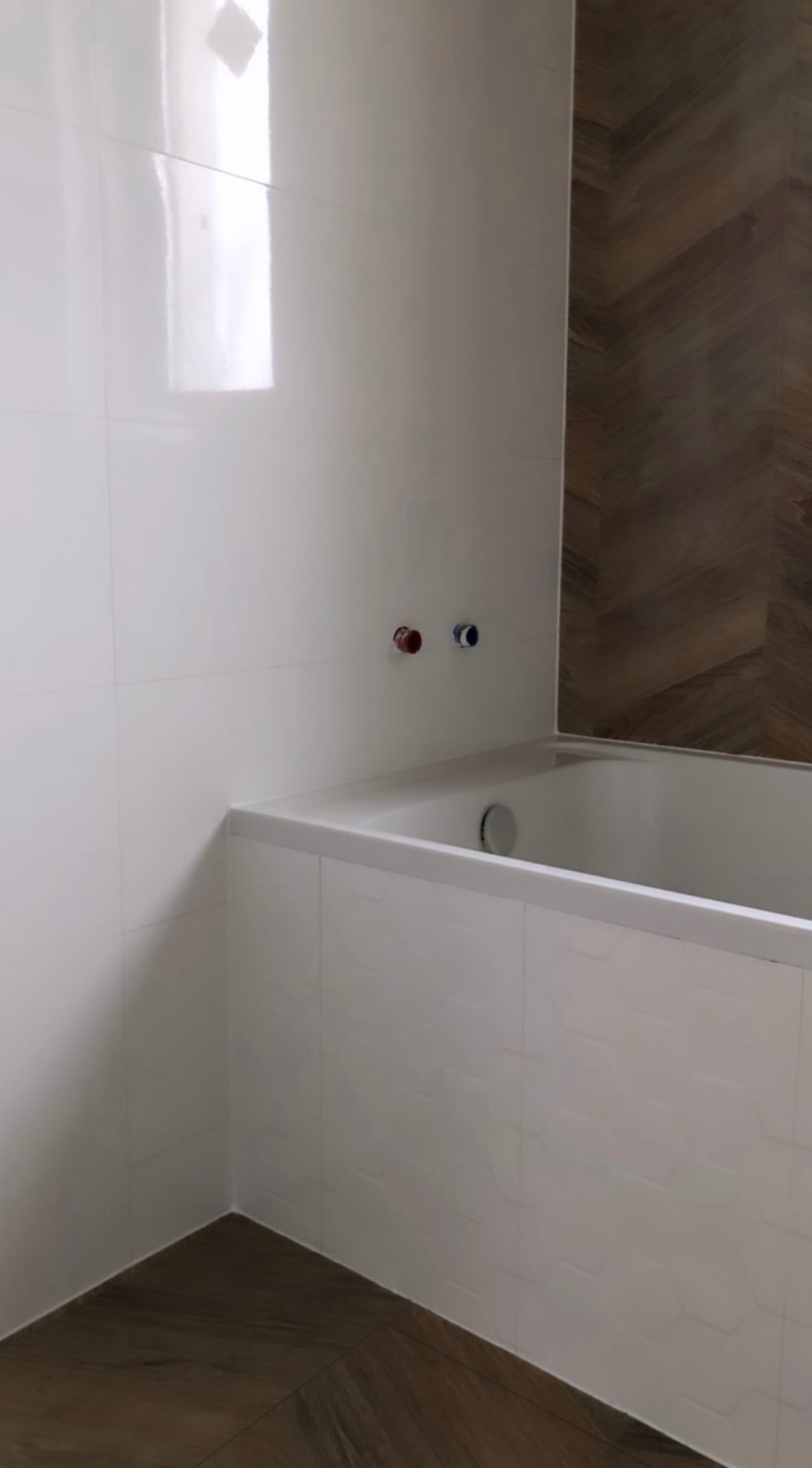 Biała wanna obudowana płytkami z delikatnym wzorem, ściana wyłożona błyszczącymi, białymi płytkami, widoczne przyłącza wodne i fragment ściany z drewnopodobnymi płytkami ułożonymi w jodełkę.