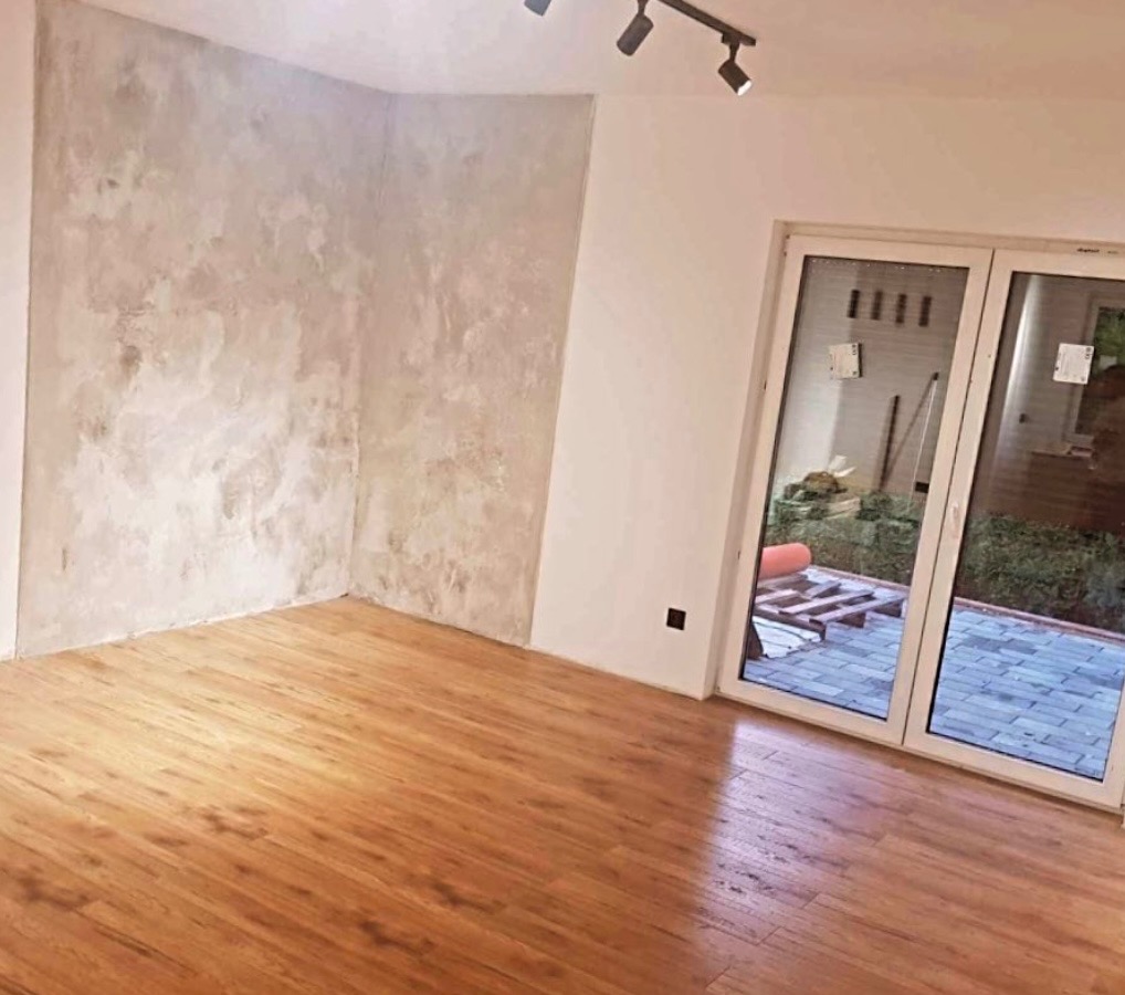 Pokój po wykończeniu: ściana z efektem dekoracyjnym w odcieniach szarości, panele podłogowe w kolorze drewna, białe okno przesuwne z widokiem na ogród.