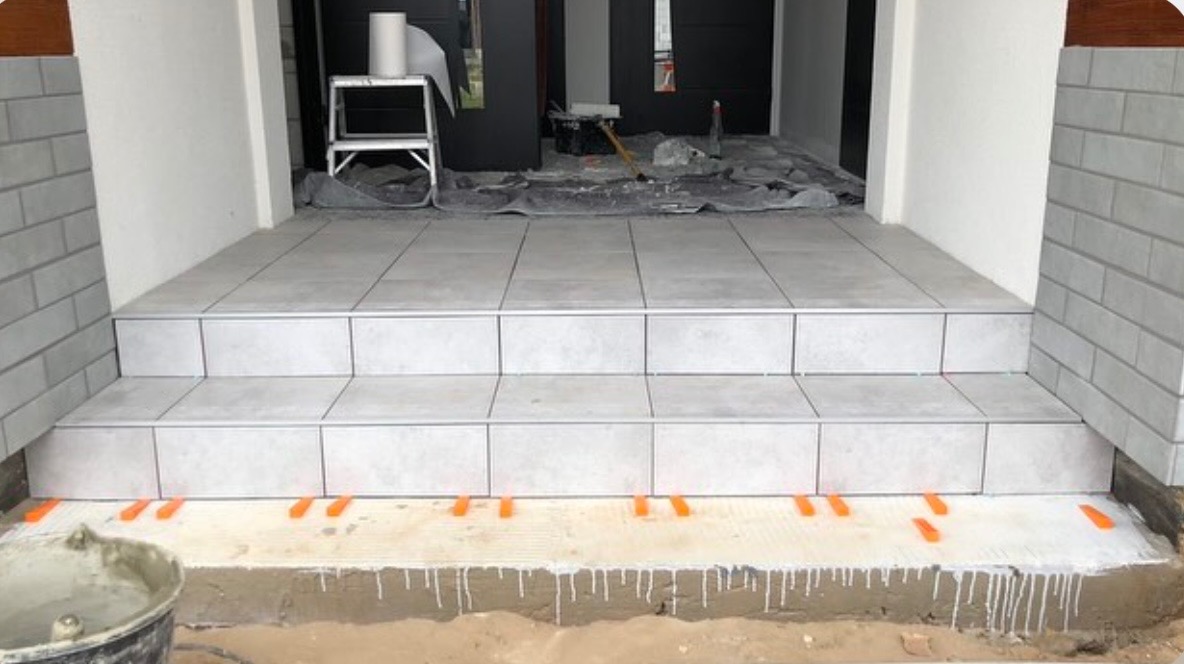 Układanie płytek na schodach wejściowych w Koninie, widoczne pomarańczowe kliny dystansowe i wiadro z zaprawą.