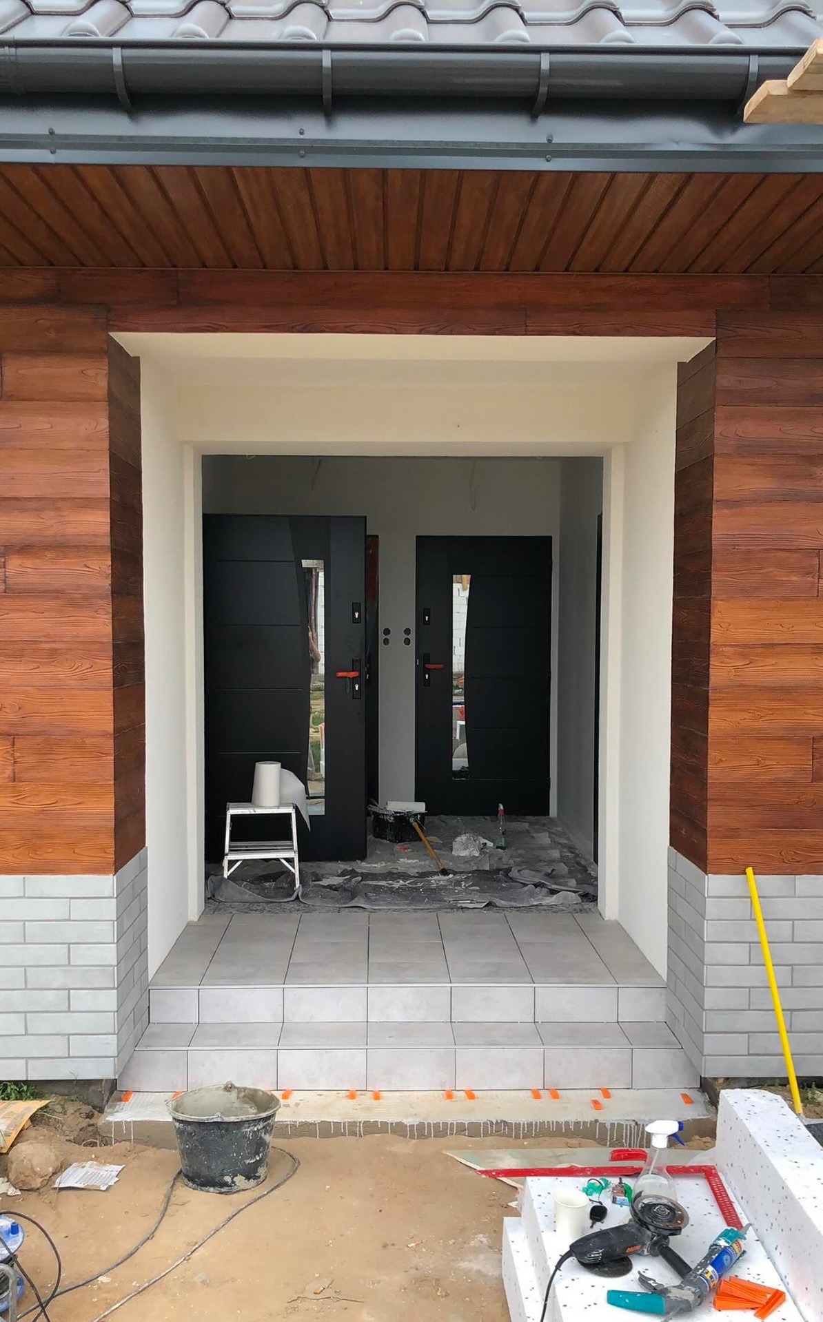 Wejście do domu w trakcie prac wykończeniowych: ułożone płytki na schodach, widoczne dwie czarne drzwi wejściowe z przeszkleniami, ściany obłożone panelami imitującymi drewno, narzędzia i materiały...