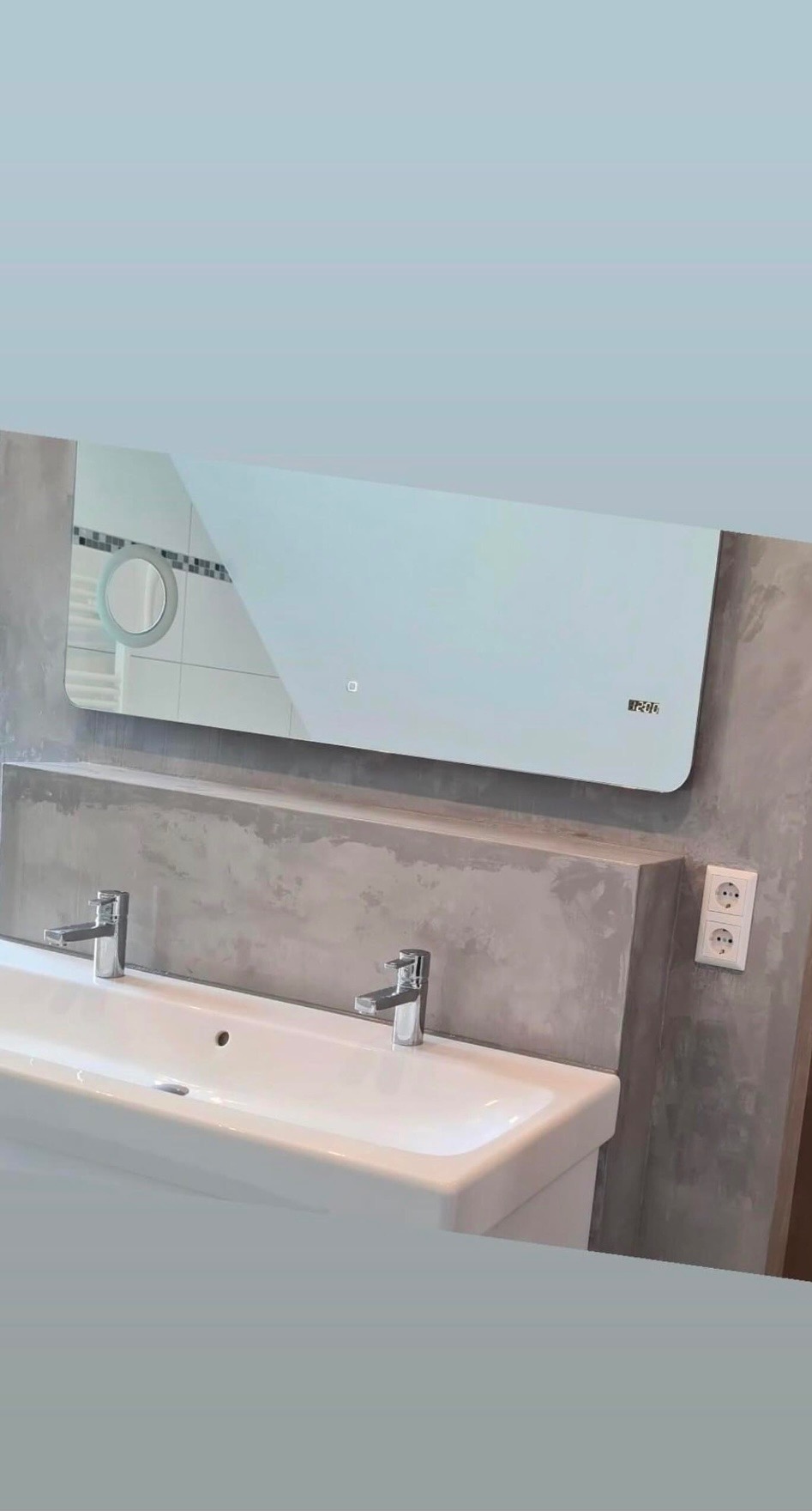 Nowoczesna łazienka z podwójną białą umywalką, chromowanymi bateriami, dużym podświetlanym lustrem z wyświetlaczem czasu i szarą ścianą z efektem betonu.