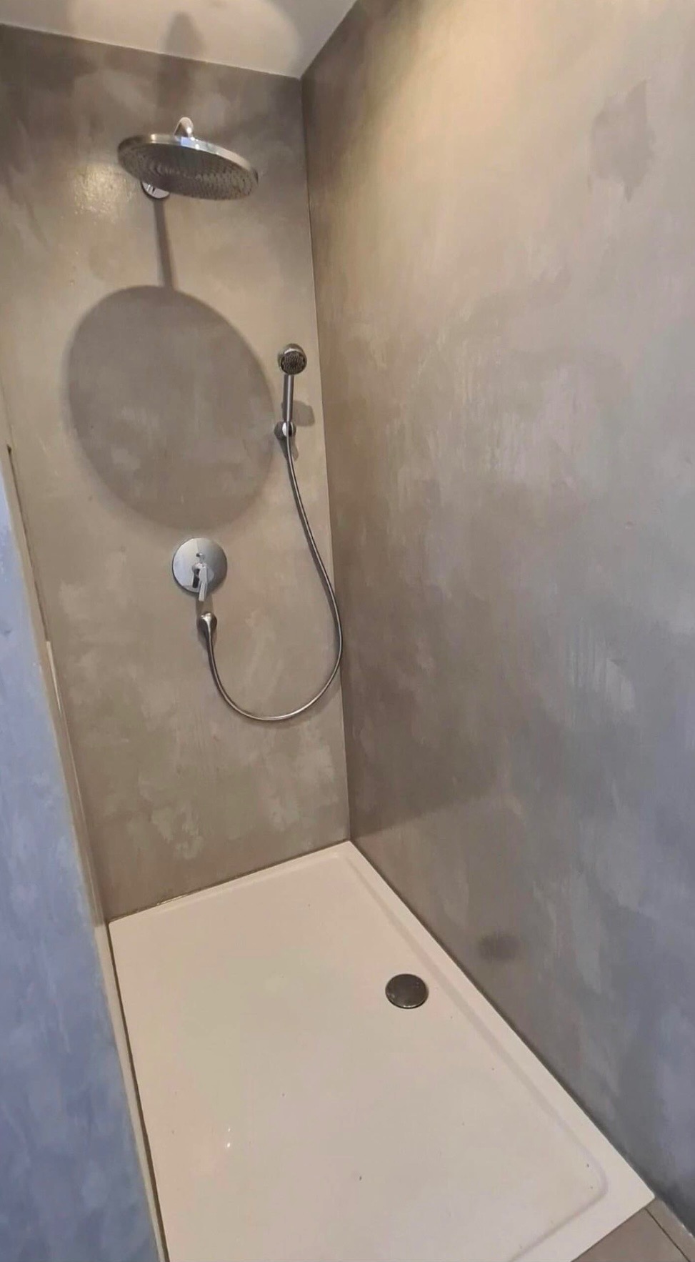 Nowoczesna kabina prysznicowa z deszczownicą i słuchawką, wykończona szarą, dekoracyjną gładzią na ścianach, z białą brodzikiem i odpływem liniowym.