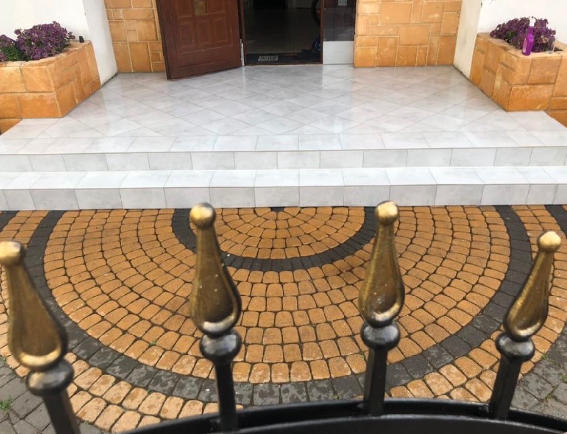 Wejście do budynku z jasnymi płytkami na schodach i okrągłym wzorem z kostki brukowej w odcieniach brązu i czerni, widoczne przez metalowe ogrodzenie z ozdobnymi elementami.