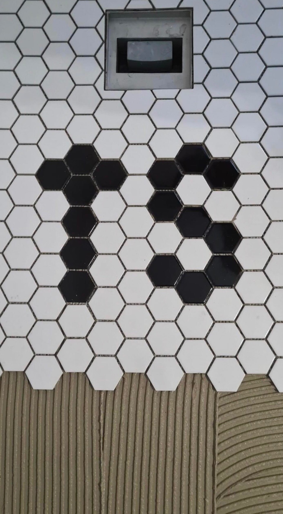 Układanie heksagonalnej mozaiki w łazience z odpływem liniowym; czarno-biały wzór na podłodze, widoczny klej do płytek.