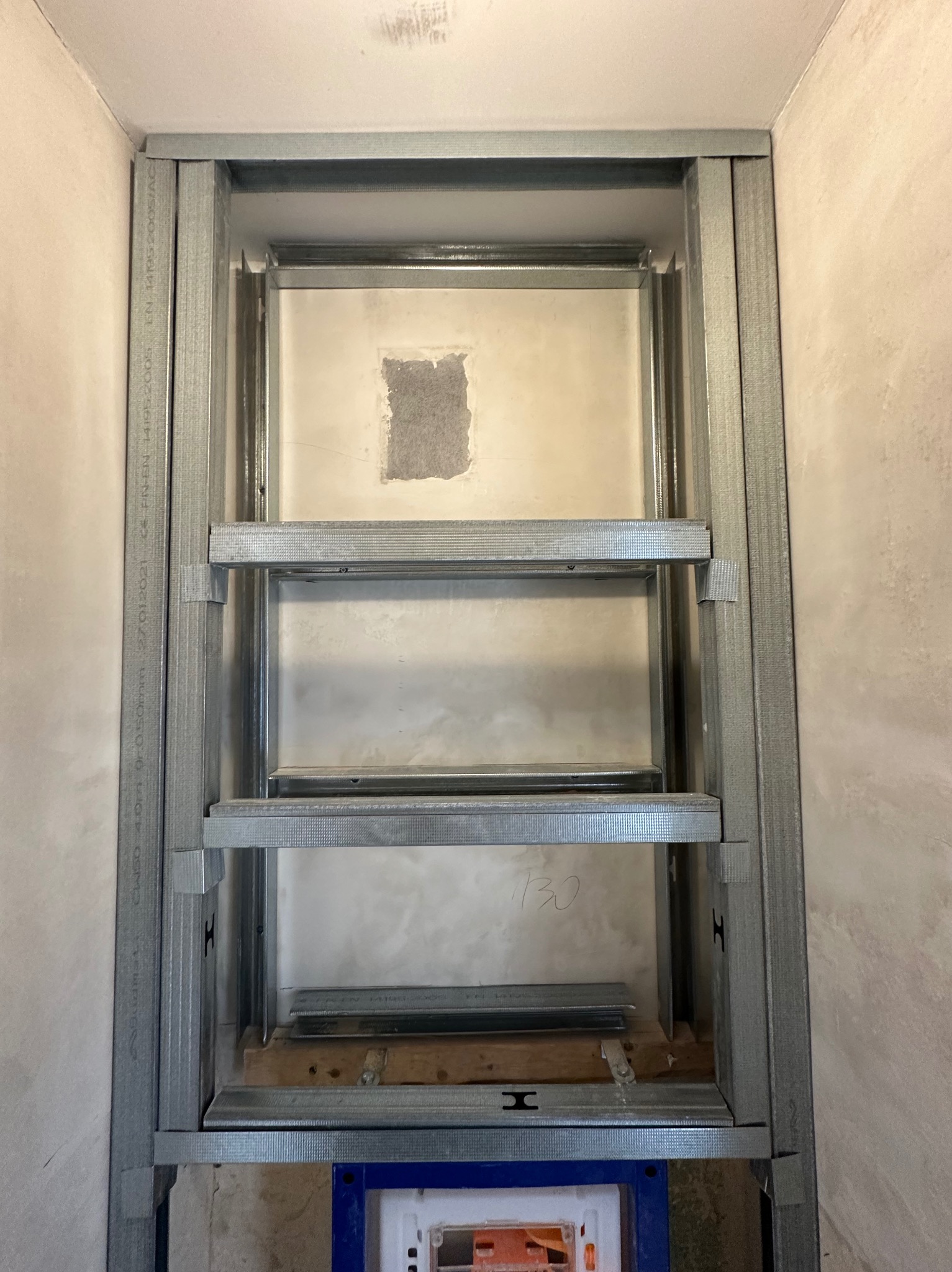 Stelaż metalowy pod zabudowę geberitu w łazience, widoczne profile, fragment ściany z niedoskonałościami i zapisaną liczbą '130', niebieski element spłuczki widoczny na dole.