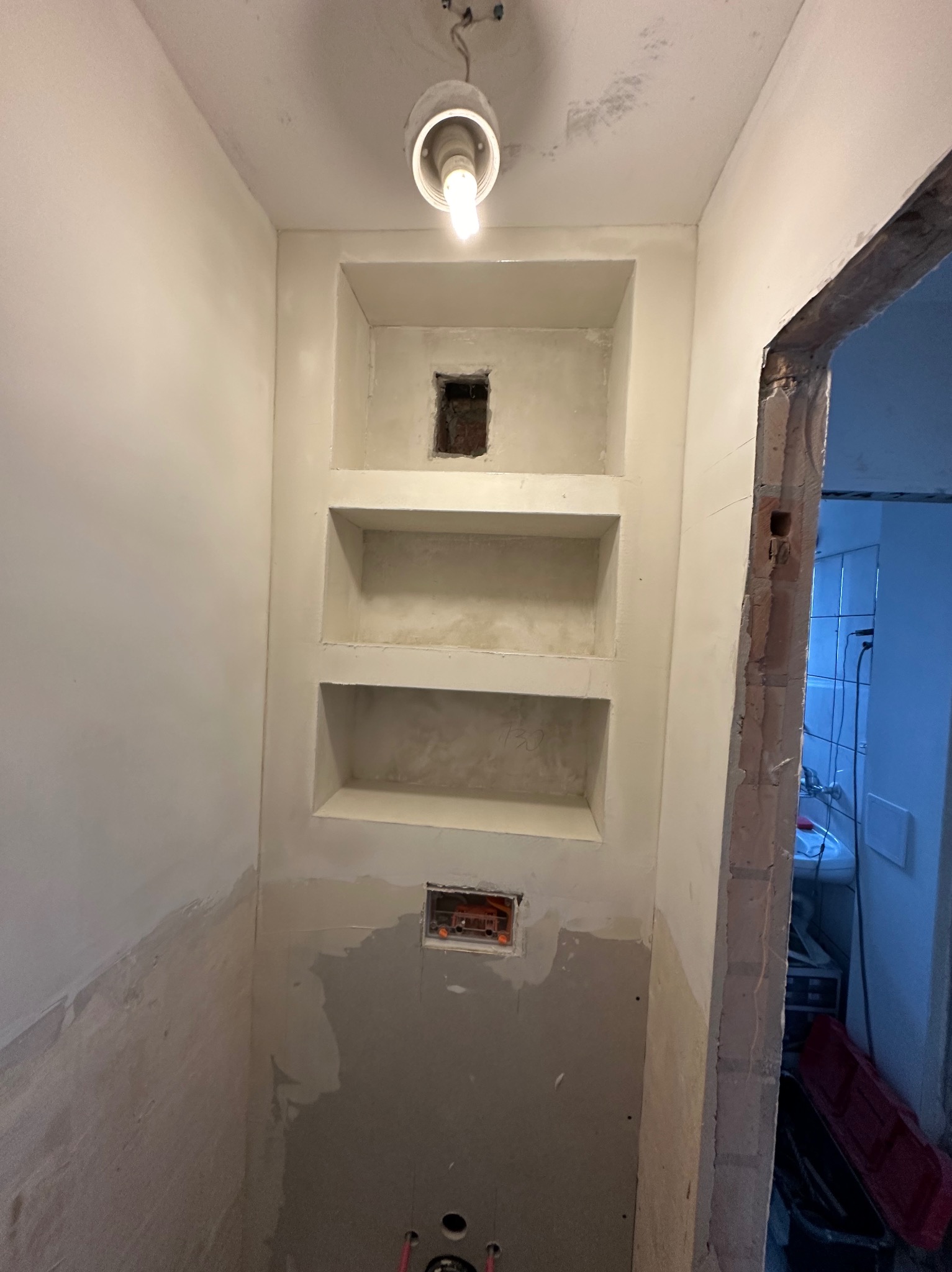 Wnęki ścienne z gładzią szpachlową w trakcie prac wykończeniowych, widoczne instalacje elektryczne i surowy otwór drzwiowy do łazienki z białymi płytkami w tle.