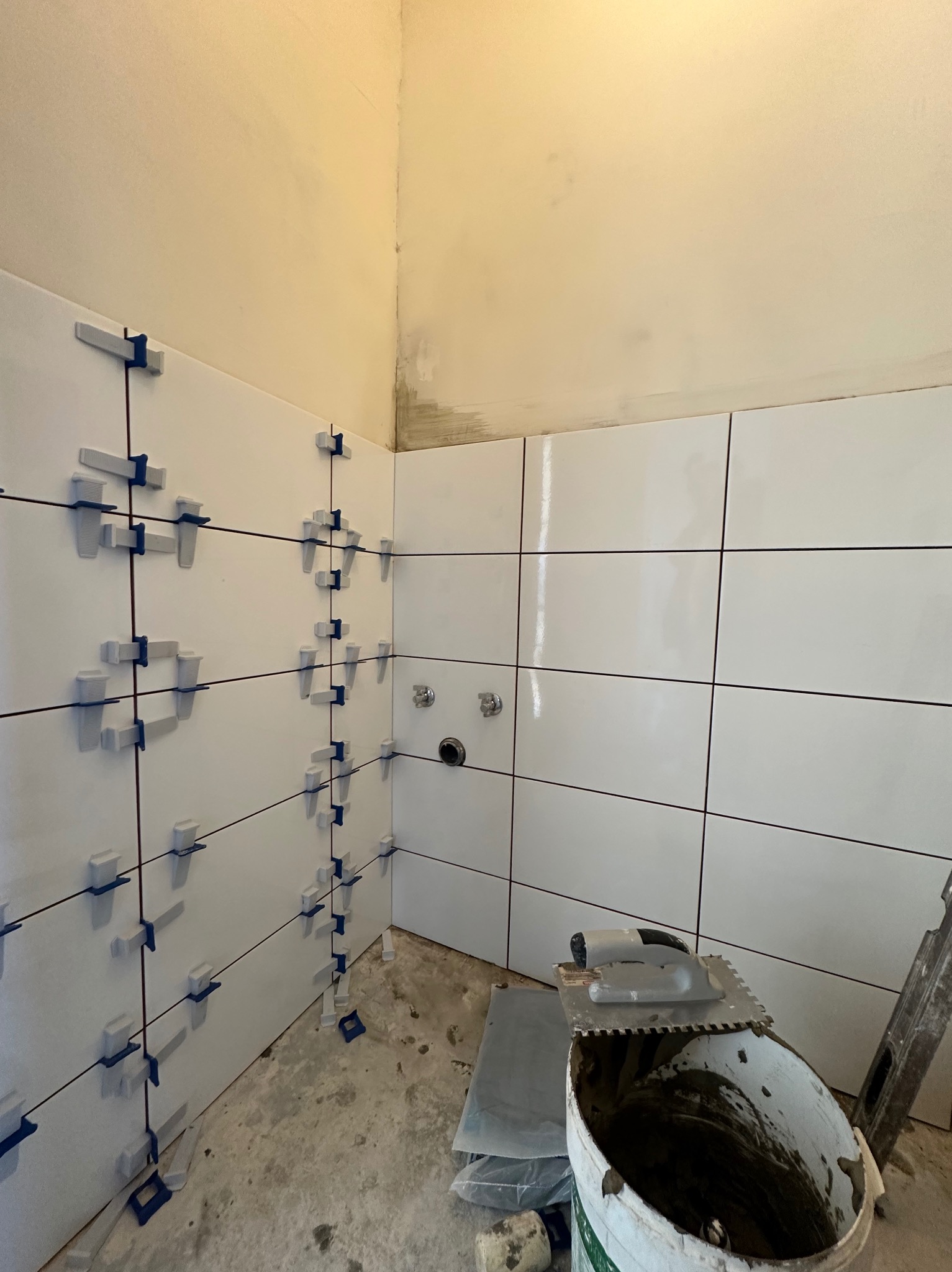 Świeżo ułożone, błyszczące, białe płytki ceramiczne na ścianie łazienki, zabezpieczone niebieskimi klipsami poziomującymi, wiadro z zaprawą i kielnia na podłodze, widoczne surowe wykończenie sufitu.