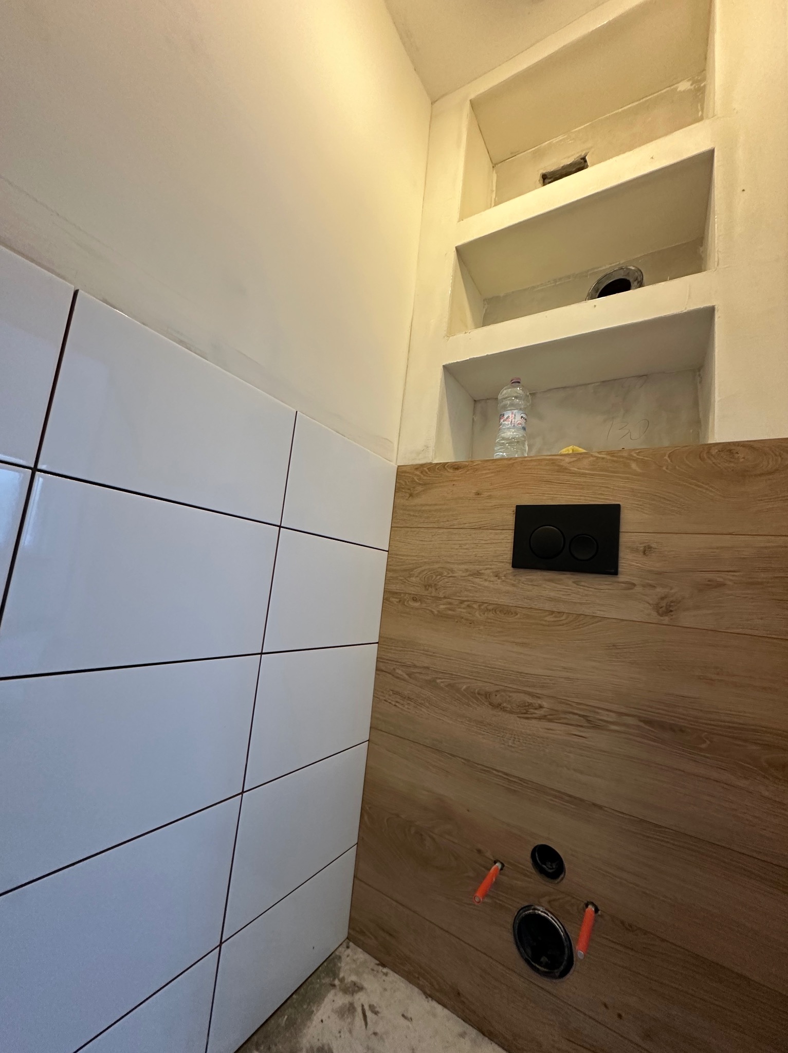 Wnęka z półkami w łazience, częściowo wykończona białymi płytkami i panelami drewnopodobnymi, widoczne otwory instalacyjne i czarny panel spłuczki, butelka wody w jednej z półek.