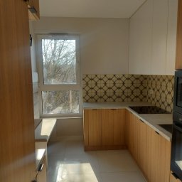 Grzegorz gzik - Jasna kuchnia z drewnianymi szafkami, wzorzystymi płytkami nad blatem i siedziskiem przy oknie z widokiem na drzewa bez liści.