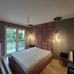 Grzegorz gzik - Sypialnia w trakcie wykańczania: łóżko z wezgłowiem z paneli tapicerowanych w odcieniach różu, wiszące lampy, grzejnik, częściowo ułożona podłoga z paneli, widok na balkon i zieleń za oknem.