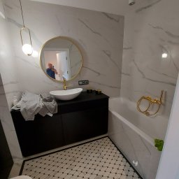 Grzegorz gzik - Nowoczesna łazienka z marmurowymi płytkami, okrągłym lustrem z złotą ramą, czarną szafką pod umywalką i mozaikową podłogą. W lustrze odbija się osoba robiąca zdjęcie.