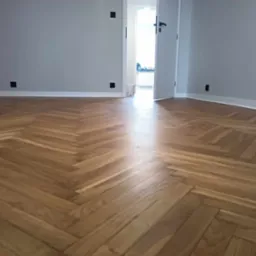 Ułożona parkietowa podłoga w jodełkę, szare ściany z czarnymi kontaktami, otwarte drzwi do jasnego pomieszczenia.