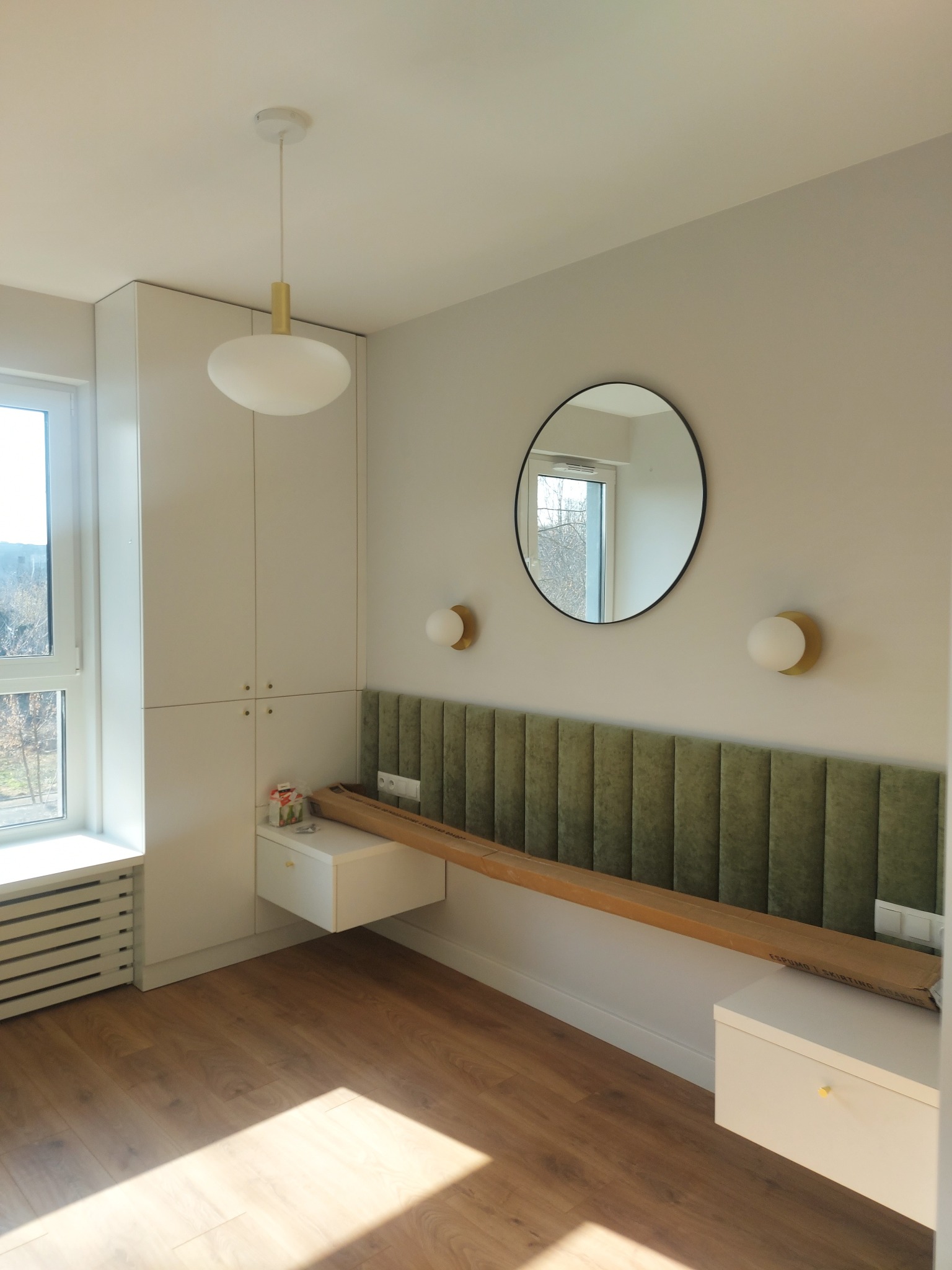 Nowoczesna łazienka w trakcie wykańczania: zielony panel dekoracyjny na ścianie, okrągłe lustro, białe szafki wiszące i lampy ścienne, podłoga z paneli, karton z listwami przypodłogowymi.