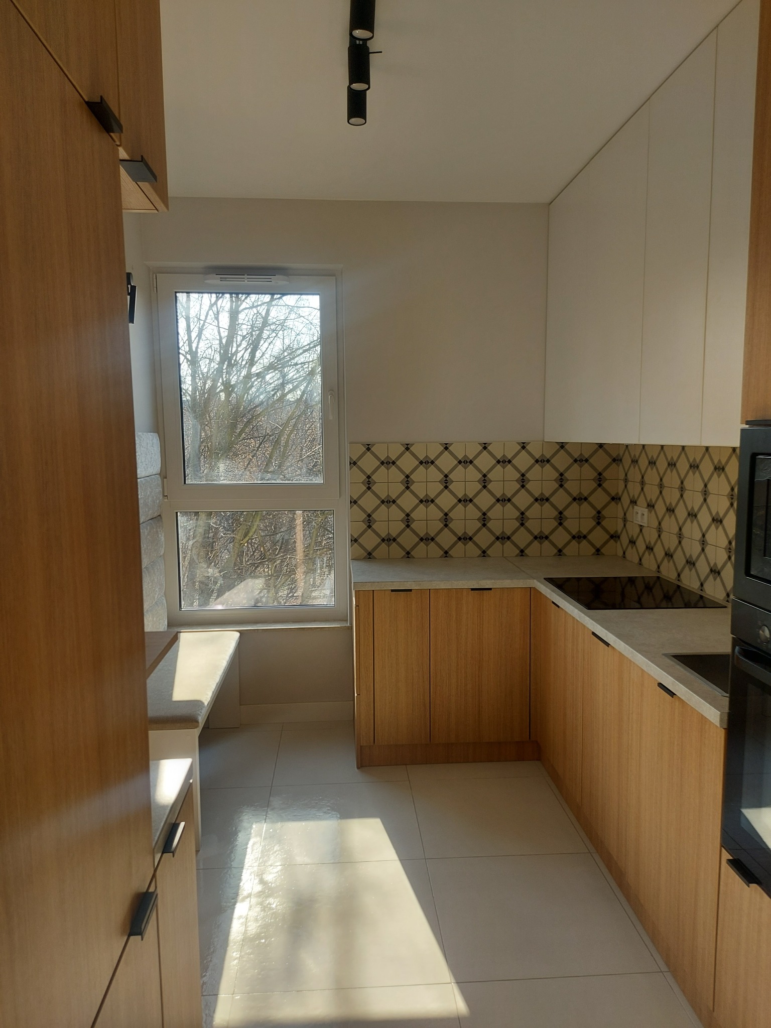 Jasna kuchnia z drewnianymi szafkami, wzorzystymi płytkami nad blatem i siedziskiem przy oknie z widokiem na drzewa bez liści.