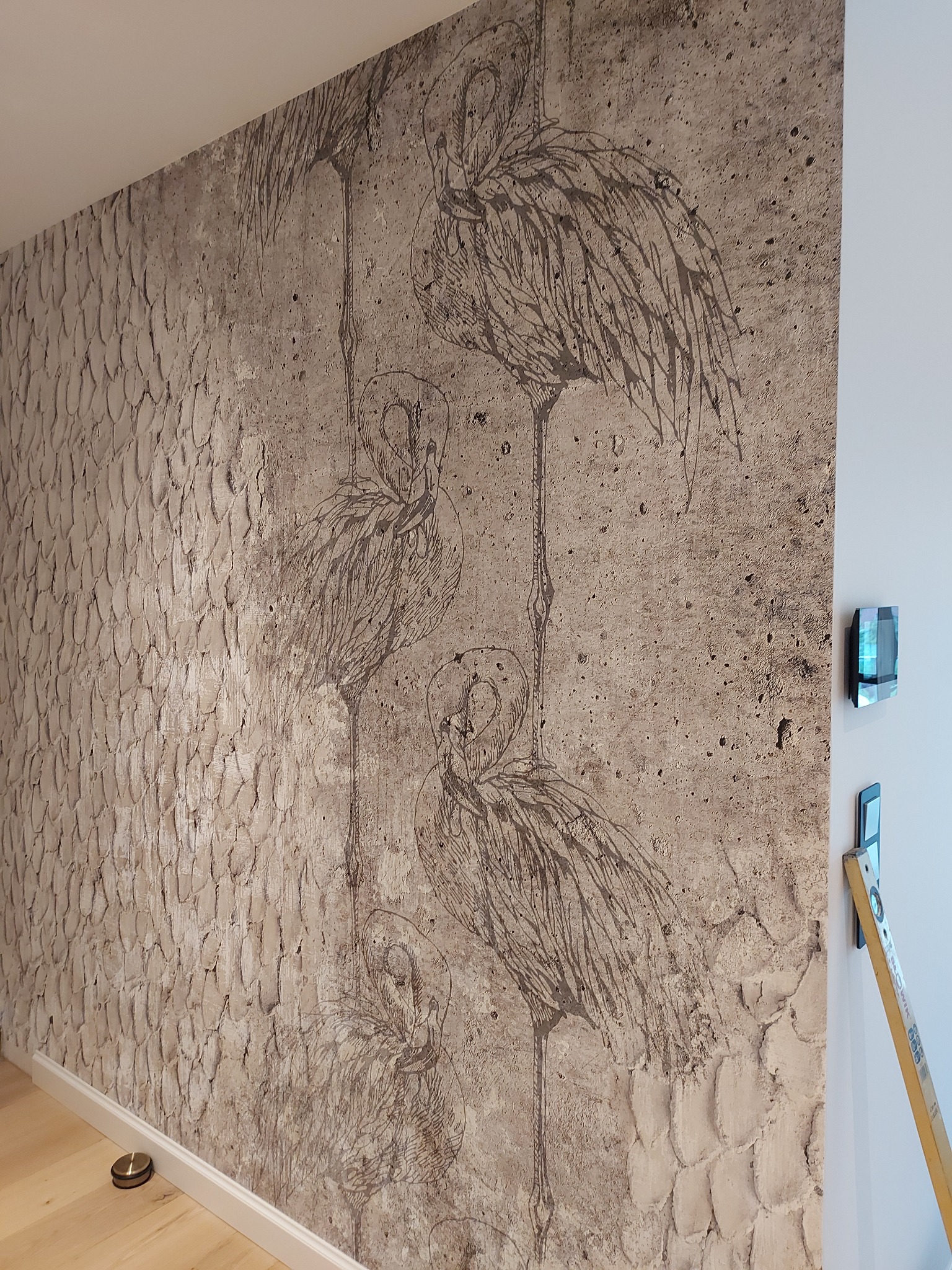 Teksturowana ściana z dekoracyjnym rysunkiem flamingów, obok pionowa poziomica i nowoczesny panel sterowania na białej ścianie.