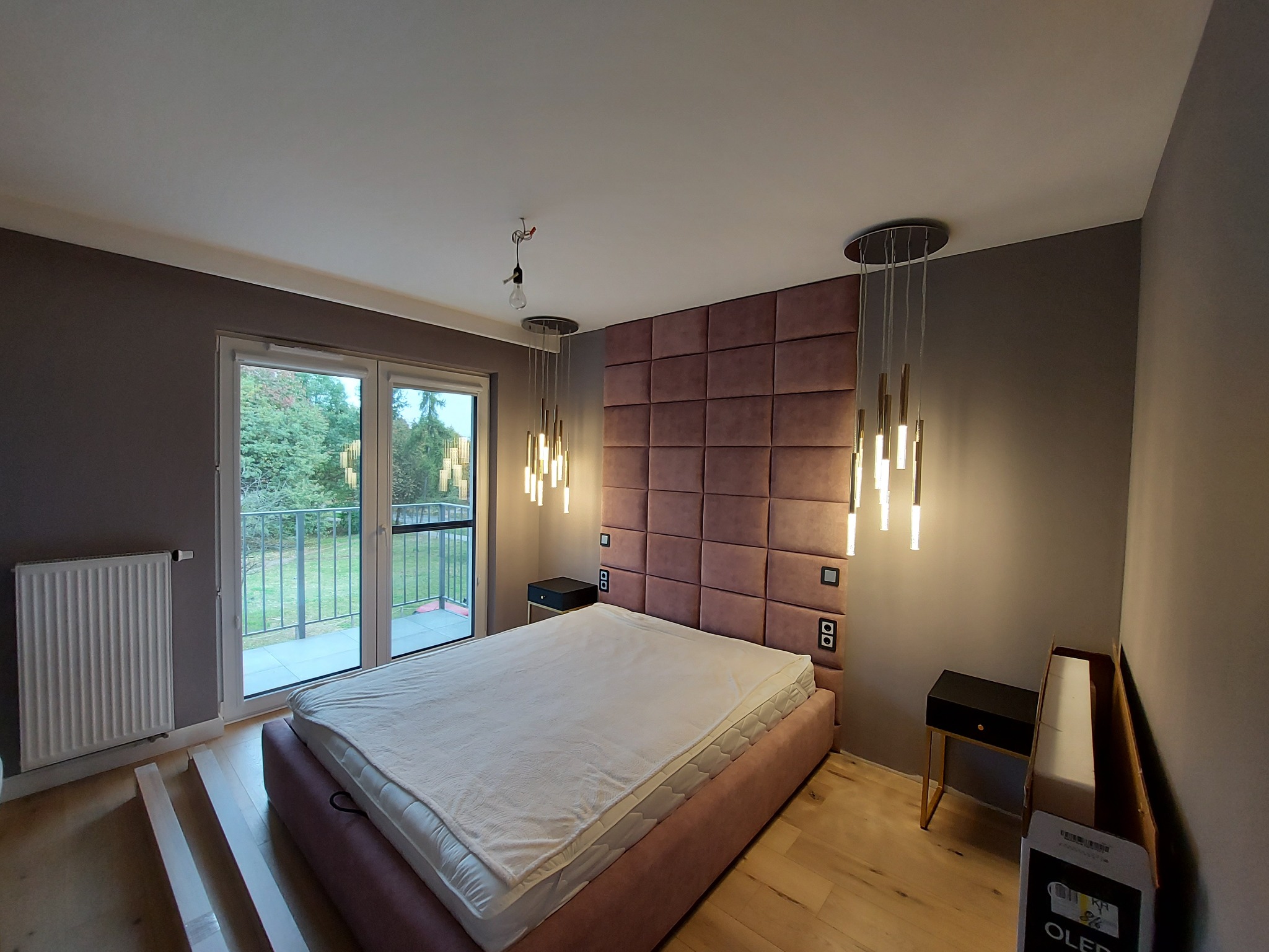 Sypialnia w trakcie wykańczania: łóżko z wezgłowiem z paneli tapicerowanych w odcieniach różu, wiszące lampy, grzejnik, częściowo ułożona podłoga z paneli, widok na balkon i zieleń za oknem.