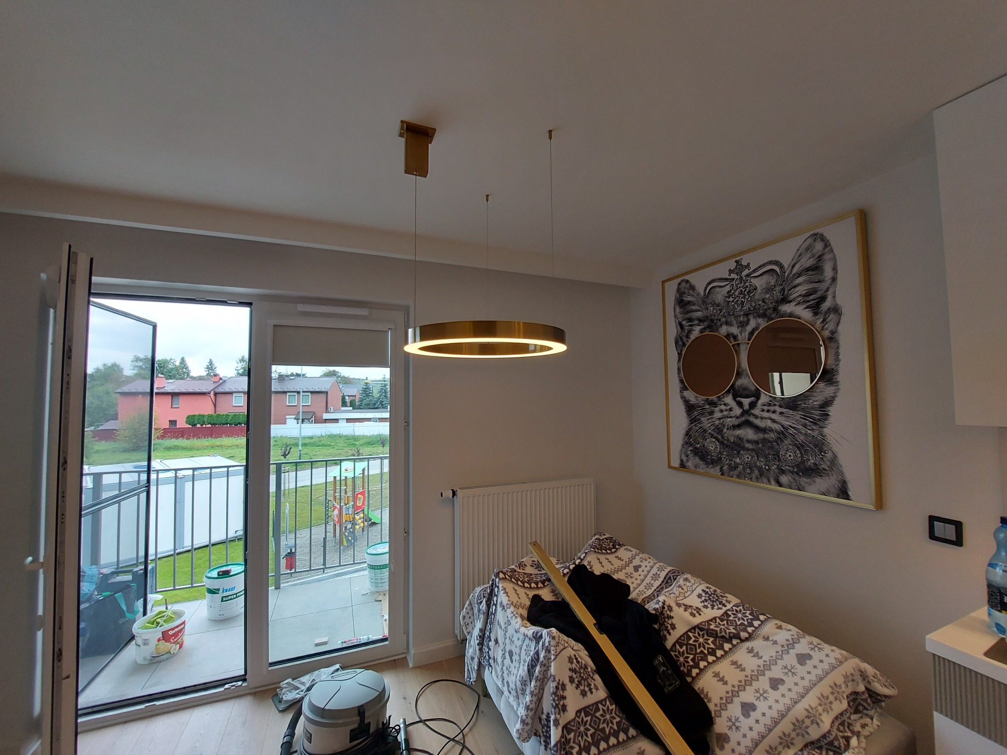 Pokój z widokiem na balkon, na podłodze narzędzia, na ścianie obraz kota w okularach, w tle widoczny plac zabaw.