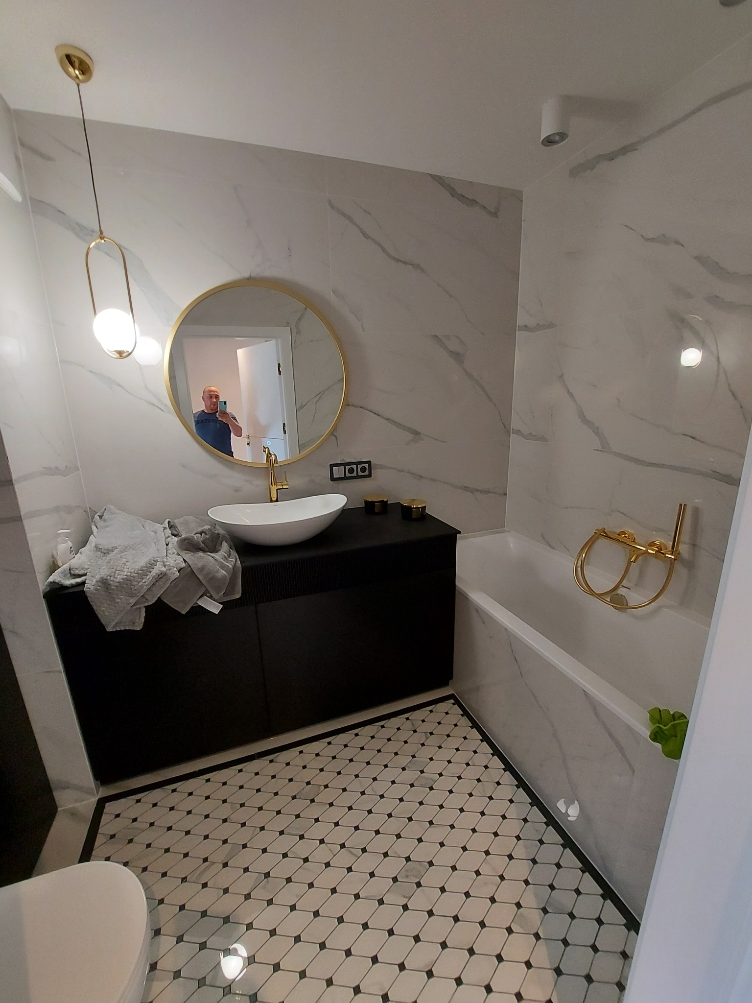 Nowoczesna łazienka z marmurowymi płytkami, okrągłym lustrem z złotą ramą, czarną szafką pod umywalką i mozaikową podłogą. W lustrze odbija się osoba robiąca zdjęcie.