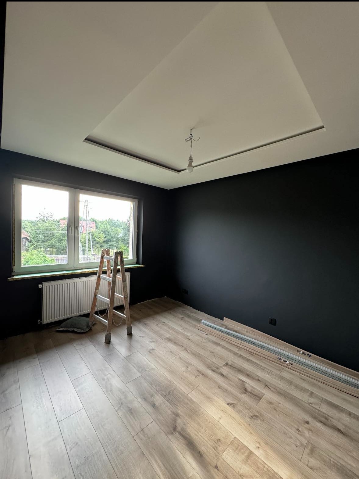 Pokój po malowaniu ścian na czarno, z drewnianą podłogą, drabiną, grzejnikiem i listwami przypodłogowymi w trakcie montażu.