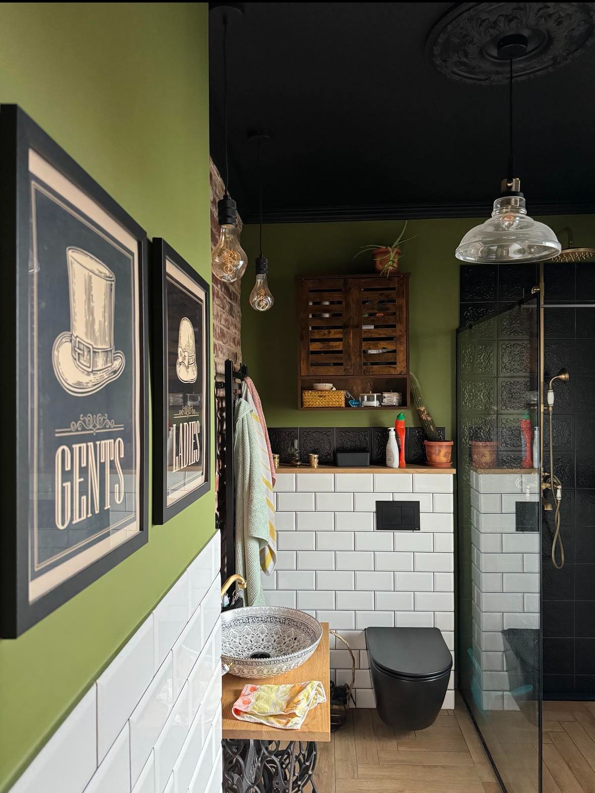 Retro łazienka z zielonymi ścianami, czarnym sufitem, kabiną prysznicową z czarnymi płytkami, umywalką na blacie z podstawą maszyny do szycia i plakatami Gents & Ladies.
