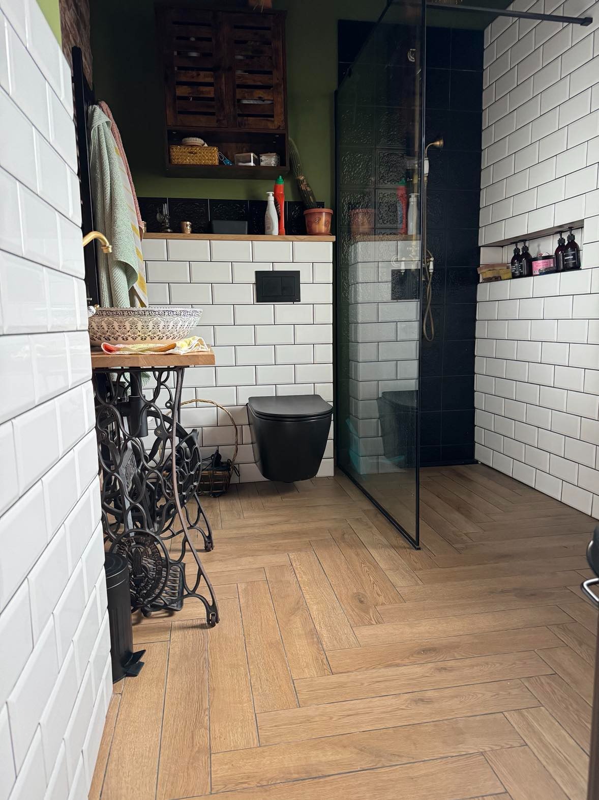 Retro łazienka z umywalką na podstawie maszyny do szycia, czarną toaletą wiszącą i prysznicem za szklaną ścianką, białe kafelki na ścianach i drewnopodobna podłoga ułożona w jodełkę.