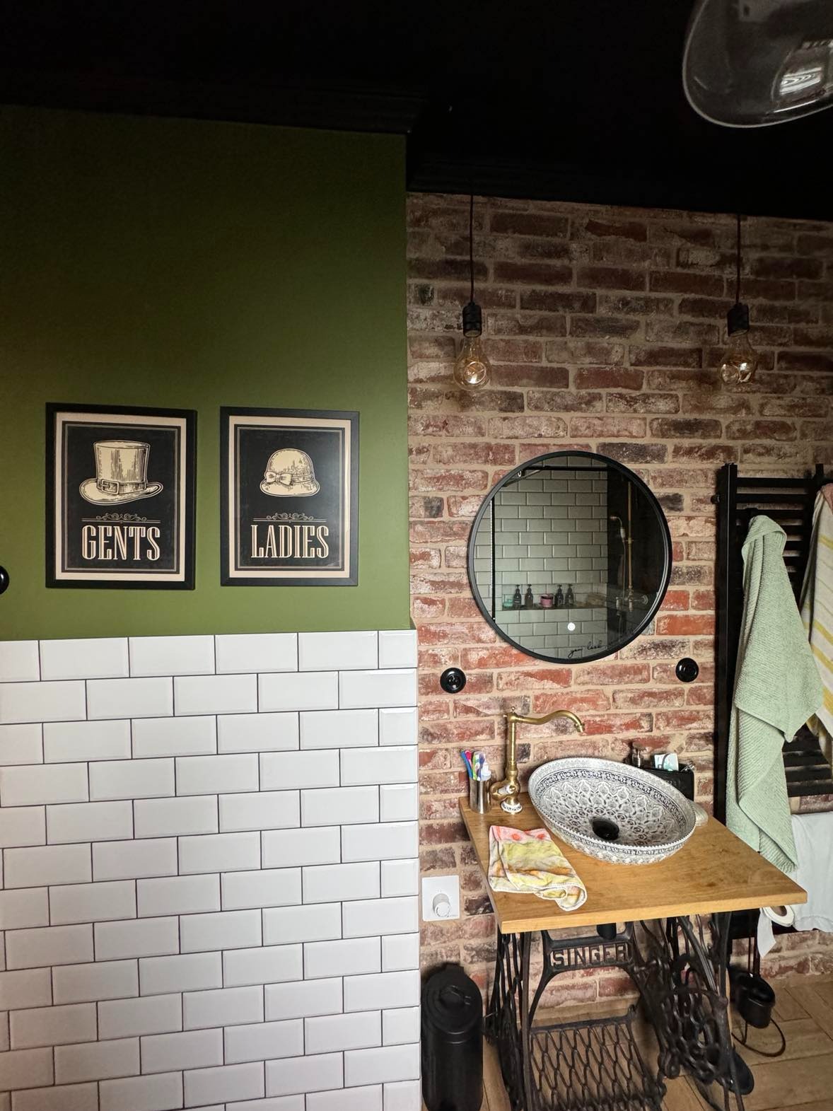 Retro łazienka z umywalką na podstawie starej maszyny do szycia Singer, ceglana ściana, białe kafelki typu metro, okrągłe lustro i plakaty z motywami damskimi i męskimi.