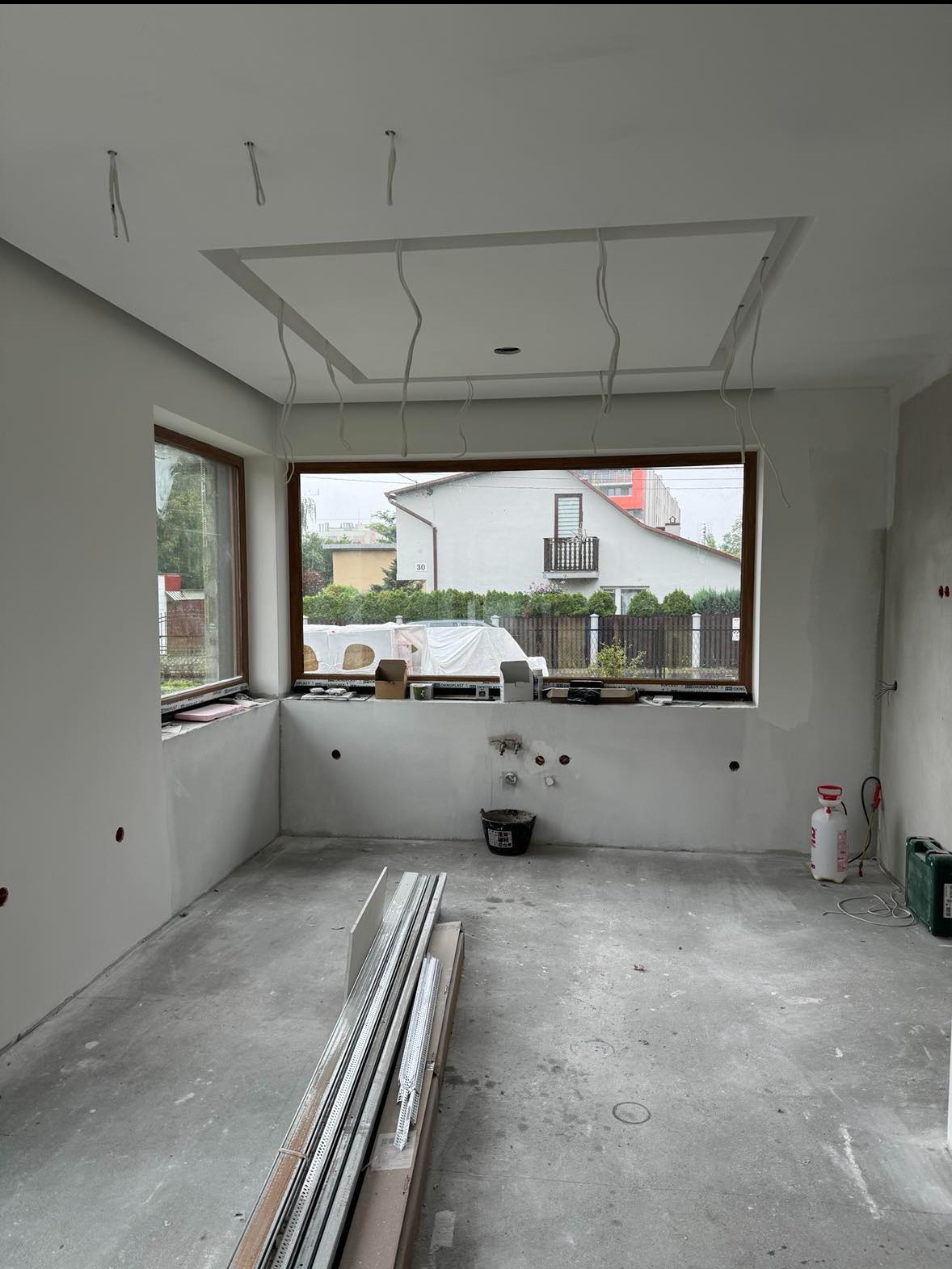 Wnętrze w trakcie prac wykończeniowych: surowy beton na podłodze, ściany przygotowane pod malowanie z widocznymi przewodami elektrycznymi, profile metalowe na podłodze, okno z widokiem na dom...